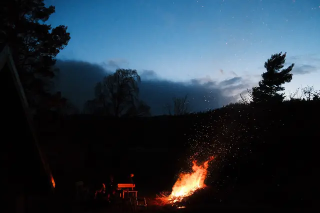 A bonfire throwing sparks into a deep blue twilight sky, with silhouetted trees and figures around the flames