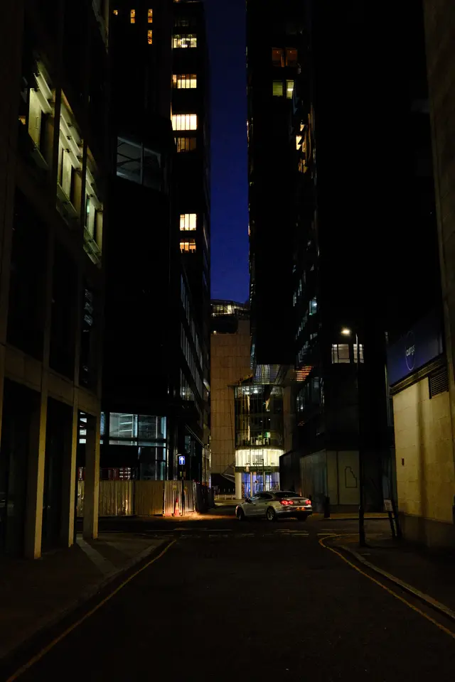 A narrow city alleyway at night, tall office buildings either side, a sliver of blue sky above