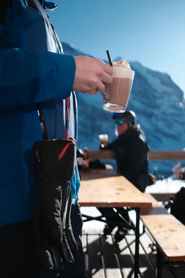 A person in ski gear holding a hot chocolate on a mountain terrace, snowy peaks behind