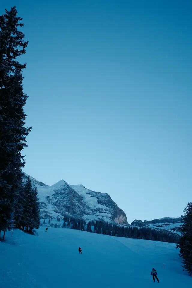 Two skiers on a wide piste at dusk with snow-covered mountains and pine trees